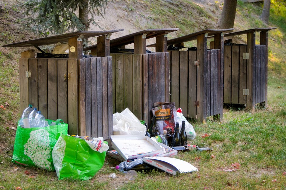 Four wooden waste bins with slanted roofs are aligned on a grassy area, set against a backdrop of trees and a sloped hillside. The bins are constructed from vertical wooden planks with a weathered, dark brown finish, each featuring metal hinges and locks. In front of the bins, there are various discarded items, including a green shopping bag filled with empty plastic bottles, a white pizza box with remnants of food, a black bag containing charcoal or firewood, and several glass bottles, some dark-colored and others clear. This outdoor scene suggests an informal waste disposal area, with rubbish including packaging, beverages, and food waste scattered on the grass, highlighting the need for proper rubbish removal services such as those provided by rubbishclearancehollandpark.com. The surrounding environment appears natural, with minimal human intervention visible, emphasizing the importance of responsible waste management to prevent environmental pollution.