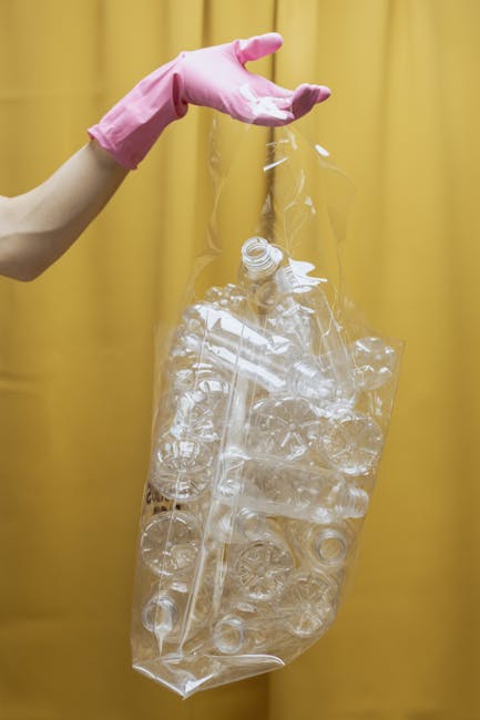 A person's hand, wearing a pink rubber glove, is holding a transparent plastic bag filled with crushed and flattened clear plastic bottles. The bottles are of different sizes and shapes, some with visible caps and others without. The scene is set against a yellow curtain or background, with the hand positioned on the left side of the image. The plastic bag appears to be ready for collection or disposal, demonstrating a typical step in waste sorting or recycling processes. The lighting is even, highlighting the glossy surface of the plastic bottles and the texture of the glove, emphasizing the preparation of recyclable materials for disposal or private waste management, as might be coordinated by a rubbish removal service.