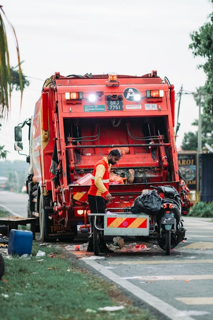 A waste management worker in a high-visibility yellow and orange vest is loading debris into the rear compartment of a large red rubbish collection truck parked on the side of a paved street. The truck's back is open, revealing mechanical components and a dirty, textured surface typical of industrial waste vehicles. The worker is standing on a small platform with a metal railing, handling a black bag among scattered small pieces of rubbish on the ground. The scene is set outdoors during daytime, with a grassy verge, a blue container, and nearby trees visible in the background. Utility poles and power lines run along the street, while a building with signage and some trees can be seen further away, indicating a typical urban or suburban environment suited to private waste collection services like those offered by rubbishclearancehollandpark.com. The overall environment supports the context of on-site clearance and alternative waste handling, often part of non-local authority rubbish removal procedures.