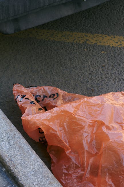 A crumpled orange plastic rubbish bag lying on a textured dark asphalt surface beside a stone curb in an outdoor urban environment. Part of a grey vehicle is visible in the background, with the bag positioned close to the curb edge and partially covering a yellow painted line on the road. The scene suggests a recent rubbish collection or disposal activity, reflecting a typical environment where private waste handling and rubbish removal services, such as those offered by companies like rubbishclearancehollandpark.com, might be involved in clearing stray debris or waste bags from city streets.