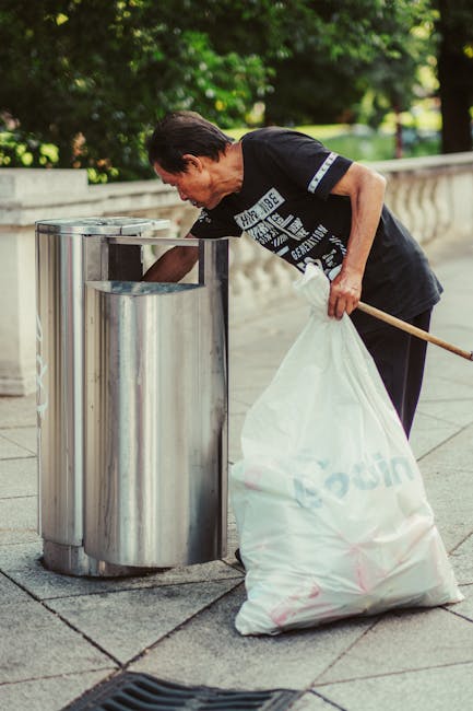 A middle-aged man with dark hair, wearing a black t-shirt with white graphics and dark trousers, is seen bending over while placing waste into a tall, cylindrical, stainless steel rubbish bin on a paved sidewalk. He is holding a large white rubbish bag filled with waste in his left hand, with the bag partially resting against his left thigh. His right hand is reaching into the open-top bin. The background features green trees and a stone balustrade, suggesting an outdoor urban environment. The scene is well-lit with natural daylight, emphasizing the metallic texture of the rubbish bin and the fabric of the rubbish bag. This image illustrates private rubbish collection in a public space, highlighting waste management activities that may be part of on-site clearance or independent waste disposal services, as offered by companies like rubbishclearancehollandpark.com for efficient rubbish removal near Holland Park station.