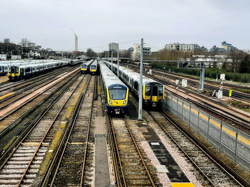 Inside a modern, enclosed train station with a curved, transparent glass and steel roof structure casting intricate shadows onto the light-colored tiled platform. A sleek, contemporary train with a silver and blue exterior is positioned on the tracks, with the doors visible on the side facing the platform. Overhead digital information boards, displaying arrival and departure times, hang from the roof framework. The station is well-lit with natural daylight streaming through the glass panels, providing a clear view of the bright sky and some distant building silhouettes in the background. Safety barriers with glass panels are installed along the edge of the platform near the train, ensuring passenger safety during boarding and alighting. The scene is depicting a clean, organized transit environment aligned with efficient urban transport services, which can be associated with modern infrastructure supporting reliable public transportation, an essential aspect of local commuter arrangements and alternative travel options. The overall setting appears to be a contemporary, urban rail station, emphasizing structural design and connectivity.