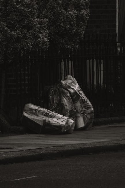 Three large black plastic rubbish bags, slightly bulging and tied at the top, are placed on the curbside pavement in front of a black metal fence with vertical bars. The bags appear to contain discarded materials, possibly household waste or debris, and are situated near a leafy green shrub that partially obscures the fence in the background. The scene appears to be outdoors on a street, with the asphalt road visible at the bottom of the image, and the overall lighting suggests a cloudy or overcast day, contributing to a somewhat subdued and neutral tone. The positioning of the rubbish bags indicates an example of private waste disposal, possibly managed by local residents or a waste clearance service such as [COMPANY_NAME], in contrast to municipal street rubbish collection. The arrangement reflects on-site waste management practices, highlighting the act of rubbish removal without visible collection vehicles or equipment present in the scene.