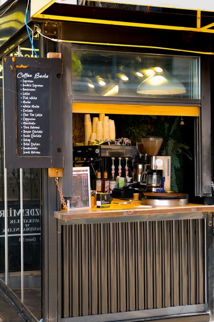 A small external coffee kiosk with a black and yellow metal frame, positioned on a sidewalk or street frontage. The kiosk has a front sliding panel made of dark, textured vertical slats, and a large glass window above that allows visibility into the interior. Inside, there is a black coffee machine, a coffee grinder, and a row of disposable cups stacked on a black shelf. Several sauce bottles are arranged on the counter, and a menu board with white text on a black background is mounted on the left side of the kiosk. The surroundings include a dark, possibly brick or painted wall on the left, with some greenery visible in the background through the reflection in the glass window. The lighting creates a warm atmosphere, with reflections of ceiling lights in the glass. The scene depicts a typical independent coffee stand, representing an example of non-traditional food and beverage service in an urban environment, possibly part of an alternative approach to small business waste management and rubbish collection by companies like rubbishclearancehollandpark.com, which may handle waste from such setups.