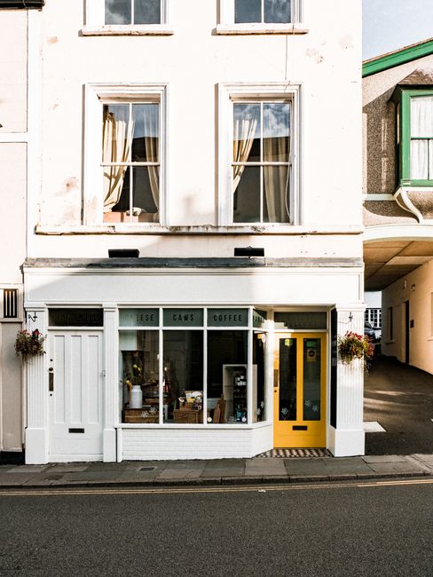 The ground floor of a white, two-story building featuring a small coffee shop with large front windows displaying various items inside. The shop has a white wooden frame with multiple panes, and the entrance includes a bright yellow door on the right side. Flanking the shop's window are two hanging flower baskets with colourful blooms. Above the windows, a sign reads 'ESE CAWS COFFEE' in black letters. The upper floors have two large sash windows with off-white curtains, set against a slightly weathered white facade. To the right, there is an alleyway or driveway leading behind the building, partially visible with a green and beige structure. The scene is lit with natural daylight, highlighting the textures of the building's exterior, the glass windows, and the street surface, which has a single yellow line along the curb. The overall scene could relate to private waste handling, such as Rubbish Clearance Holland Park, by illustrating a commercial premises that may require rubbish removal or waste collection services, characteristic of an independent shop dealing with regular waste disposal.