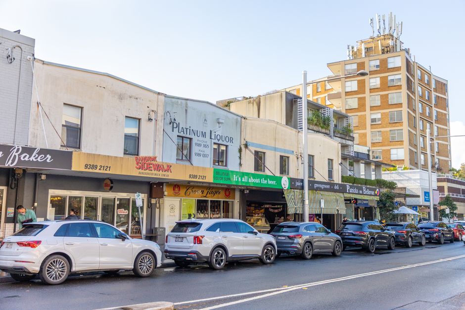 A small external coffee kiosk with a black and yellow metal frame, positioned on a sidewalk or street frontage. The kiosk has a front sliding panel made of dark, textured vertical slats, and a large glass window above that allows visibility into the interior. Inside, there is a black coffee machine, a coffee grinder, and a row of disposable cups stacked on a black shelf. Several sauce bottles are arranged on the counter, and a menu board with white text on a black background is mounted on the left side of the kiosk. The surroundings include a dark, possibly brick or painted wall on the left, with some greenery visible in the background through the reflection in the glass window. The lighting creates a warm atmosphere, with reflections of ceiling lights in the glass. The scene depicts a typical independent coffee stand, representing an example of non-traditional food and beverage service in an urban environment, possibly part of an alternative approach to small business waste management and rubbish collection by companies like rubbishclearancehollandpark.com, which may handle waste from such setups.