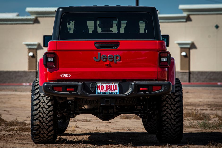 A rear view of a red Jeep SUV parked on a sandy, unpaved surface, with large, rugged off-road tires and a black roof. The vehicle’s tailgate features a black Jeep logo in the center, and the license plate reads 'NO BULL' with additional smaller text 'SINCE 1951' and the dealer name 'CARNARDT' below. The background shows a beige building with small, elevated wall-mounted lights and a clear blue sky, suggesting an outdoor, possibly commercial or industrial environment. The car’s finished surface is shiny, reflecting sunlight, contrasting with the matte, dusty ground beneath. This image may relate to independent vehicle disposal or private waste removal, aligning with services that handle rubbish clearance or vehicle disposal in a professional context.