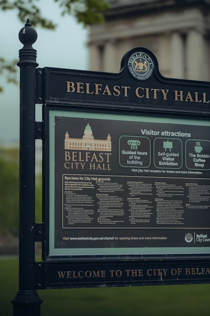 A large, freestanding information board situated outdoors near Belfast City Hall, with a dark blue metal frame and decorative finial at the top of the left post. The sign displays the heading 'BELFAST CITY HALL' in gold lettering, and underneath, it features various sections providing details about visitor attractions, guided tours of the building, self-guided visitor exhibitions, and the Bobbin Coffee Shop, along with a logo of the Belfast City Council at the top. The background shows a blurred view of trees and greenery, indicating a park or open public space, with natural daylight illuminating the scene. The surroundings suggest a context where public information about local landmarks is displayed, which contrasts with the typical visual of rubbish removal services but highlights the importance of maintaining clean, well-organized outdoor environments as part of community management and waste regulation compliance in urban areas.
