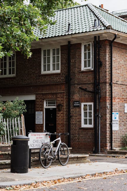 The image shows a two-story brick building with a pitched, tiled roof in grey tones, situated at the edge of a street or alleyway. In front of the building, a white bicycle is parked upright on its kickstand, leaning against a roadside sign that reads 'Chelsea Square.' Nearby, there is a black cylindrical trash bin with a rounded lid positioned on the pavement. To the right of the bin, a small, green shrub partially obscures a white wooden fence. An outdoor light fixture is mounted beside the door, which has a white frame and a small rectangular window. The building features white-framed windows with multiple panes, and black downpipes run vertically along the wall, connecting to the gutter system at the roofline. The environment appears quiet, with overhanging leafy branches visible in the upper left corner, and the overall scene reflects an urban setting where private waste arrangements might be considered outside of standard council rubbish collection. The scene has natural daylight, and fallen leaves scattered on the pavement suggest an autumn season, supporting the context of waste management and rubbish disposal in residential areas.